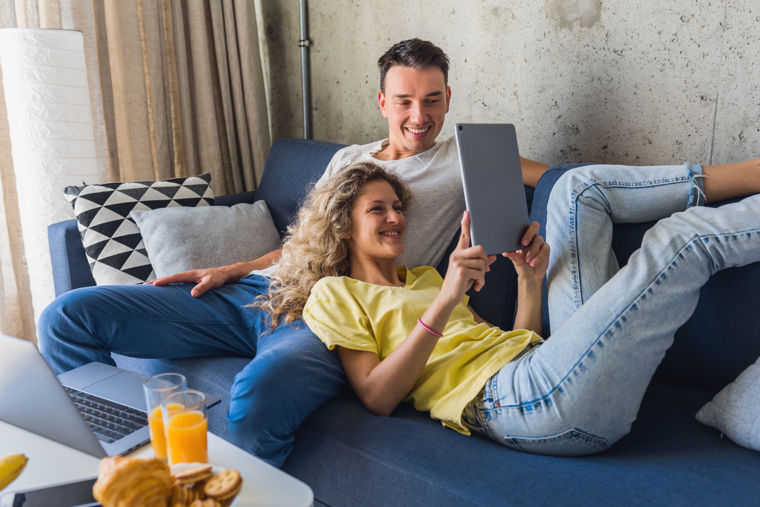 Young,Couple,Sitting,On,Sofa,At,Home,Looking,In,Tablet